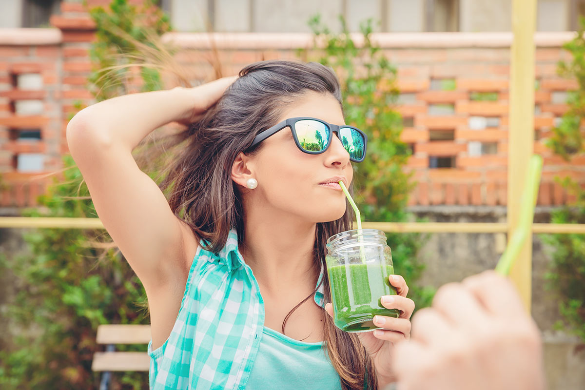 Młoda kobieta w okularach przeciwsłonecznych pije zielony sok warzywny przez słomkę. 
